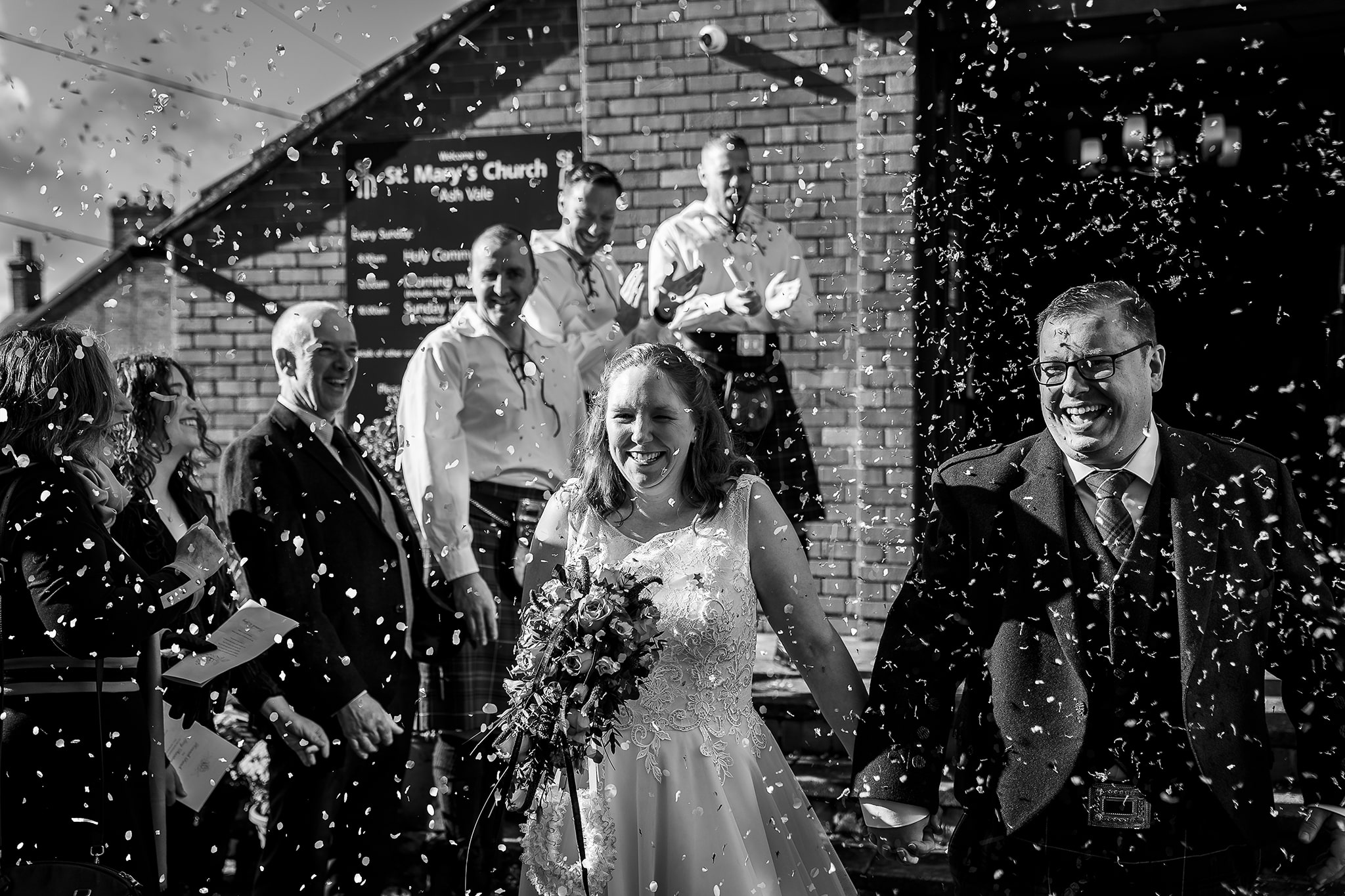 Black and white photo of wedding couple walking through lots of confetti at St Mary’s Church in Ash Vale by Emma Seaney